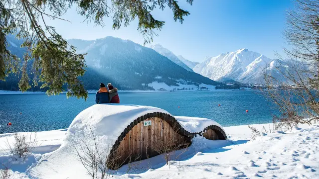 Zwei Personen blicken über einen blauen See auf verschneite Berge.